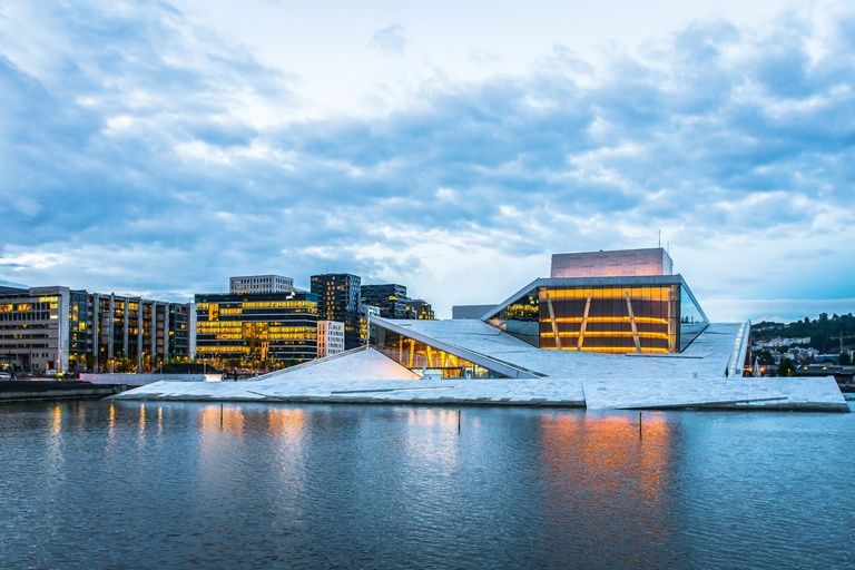 The Oslo Opera House and the Bjørvika waterfront lit at dusk, reflected in the fjord